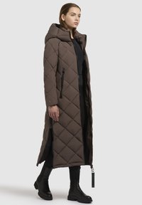 Kvinde iført en lang, brun quiltet hættejakke med sorte lynlåsdetaljer, sort tøj og sorte klodsede støvler foran en hvid baggrund.