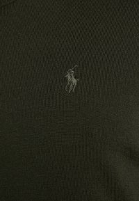 Olivově zelený pletený svetr s žebrovanou texturou, zdobený tónovaně vyšívaným logem na hrudi. Klasický kulatý výstřih, minimalistický design.