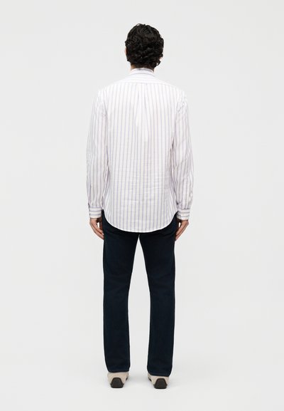 Άνδρας με σκούρα, σγουρά μαλλιά που φοράει λευκό ριγέ πουκάμισο με μακριά μανίκια, σκούρο παντελόνι και ανοιχτόχρωμα παπούτσια, στέκεται με την πλάτη προς τα εμπρός.