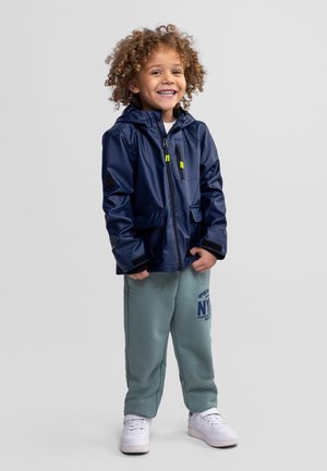 Niño sonriente con cabello rizado, vestido con una chaqueta impermeable azul marino, pantalones deportivos grises con texto azul y zapatillas blancas, de pie sobre un fondo gris.