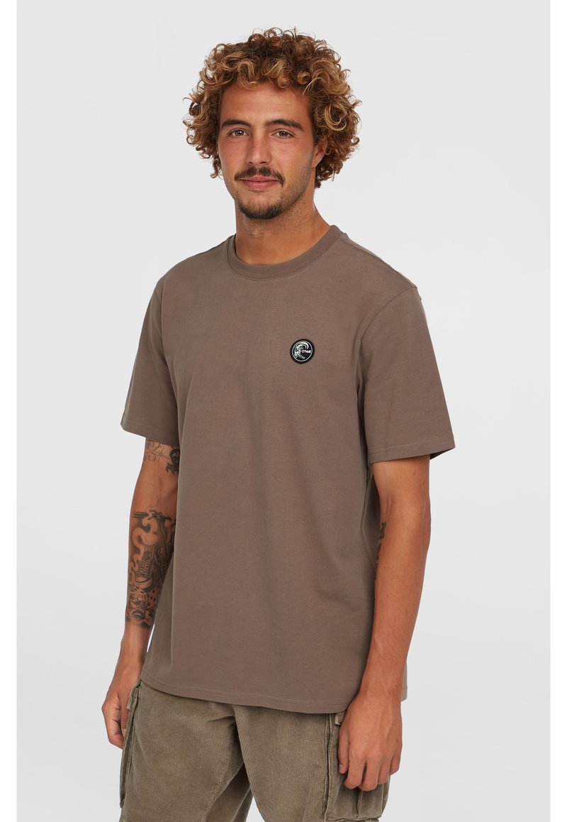 Brun bomulds t-shirt med rund halsudskæring, korte ærmer og et sort cirkulært logo på brystet. Modellen bærer lysebeige cargo bukser.