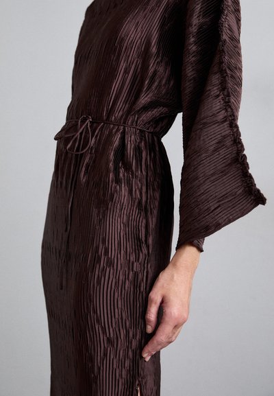 Person wearing a dark brown pleated dress with wide sleeves and a thin waist tie, hand relaxed by their side against a plain background.