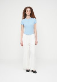 Νέα γυναίκα με κατσαρά μαλλιά, φορώντας ανοιχτό μπλε κοντομάνικο πουκάμισο, λευκό παντελόνι με φαρδύ πόδι και μαύρα μπαλαρίνες, στέκεται μπροστά σε λευκό φόντο.