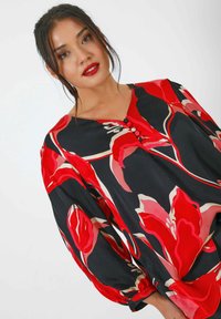 Zwarte blouse met groot rood bloemenpatroon, heeft een V-halslijn, drie knopen en luchtige mouwen met gathered manchetten.