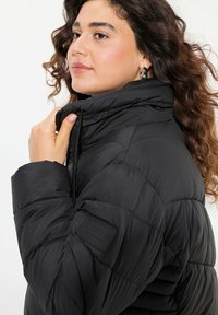 Svart puffjacka med hög krage, med quiltat mönster, dragkedjestängning och sidofickor. Läderpatch på ärmen ger extra detalj.