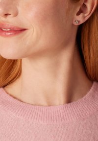 Boucles d'oreilles en argent avec un design en spirale, portées par une personne à la peau claire et aux cheveux roux, complétant un pull rose avec un col côtelé.