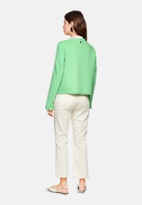 Mintgrüner, taillierter Blazer mit langen Ärmeln, der fransige Kanten aufweist, kombiniert mit cremefarbenen, geraden Hosen und hellen Sandalen.