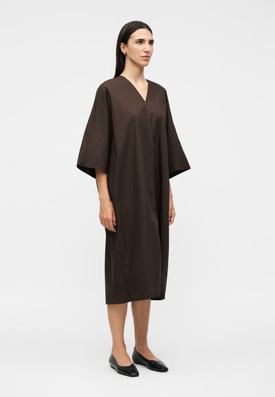 Γυναίκα με ίσια σκούρα μαλλιά που φοράει ένα άνετο, σκούρο καφέ φόρεμα μέχρι τα γόνατα με φαρδιά μανίκια και μαύρα επίπεδα παπούτσια, στέκεται μπροστά σε λευκό φόντο.