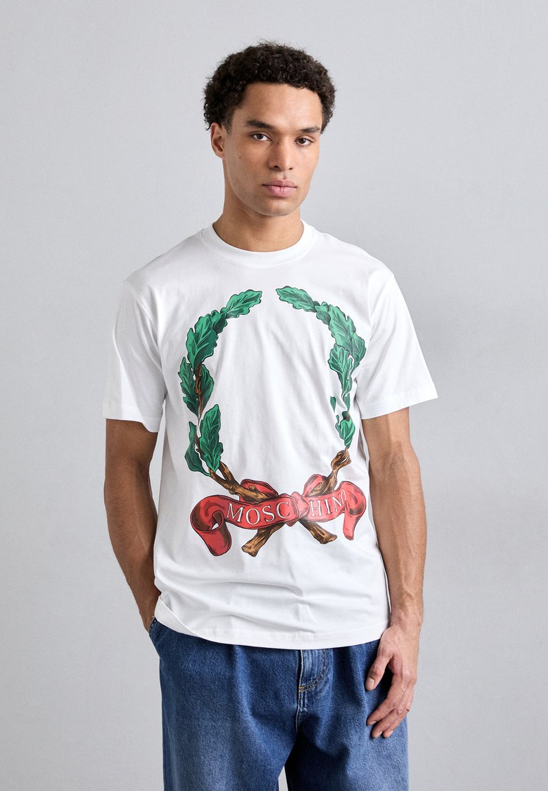 White cotton t-shirt featuring a green laurel wreath graphic and a red ribbon with "MOSCHINO" text at the bottom. Short sleeves, classic fit.