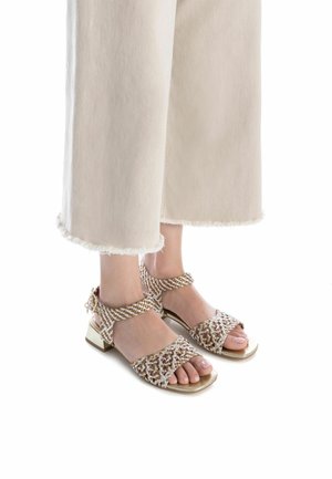 Sandalias beige con correas entrelazadas, un pequeño tacón cuadrado y acentos dorados. El diseño presenta un patrón floral en la parte superior.