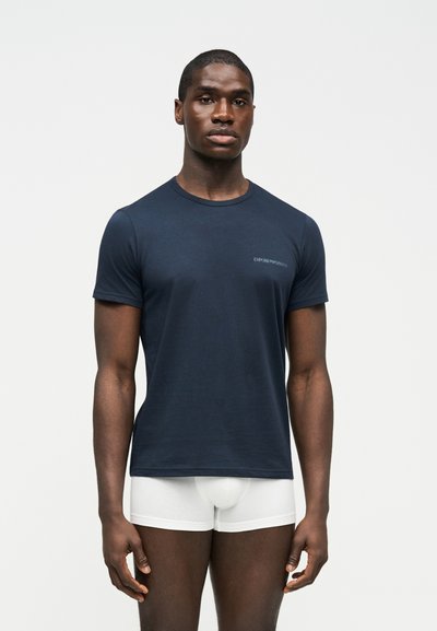 Νέος άνδρας που στέκεται προς τα εμπρός, φορώντας μπλε σκούρο t-shirt Emporio Armani και λευκά ανδρικά εσώρουχα τύπου boxer, μπροστά από απλό φόντο.