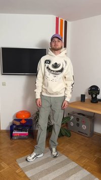 Felpa con cappuccio crema con design grafico e dettagli neri, pantaloni in velluto a coste verde e sneakers bianche chunky. In piedi su un tappeto a righe.