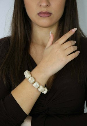 Beiges Keramikarmband mit glänzendem Finish, das sechs abgerundete Perlen und goldene Akzente aufweist, getragen an einem teilweise sichtbaren Arm.