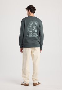 Felpa verde con stampa grafica sulla schiena, che presenta il testo "ELEVATE ADVENTURE" e un design stilizzato. Abbinata a pantaloni di colore crema.