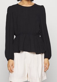 Top peplum nero con maniche lunghe a sbuffo, vita arricciata e texture morbida, abbinato a pantaloni corti di colore chiaro.