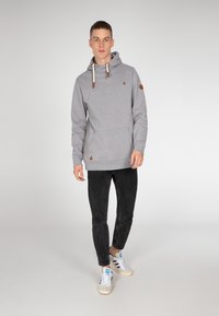 Grijze hoodie met trekkoord, leren accenten en zijzak, gecombineerd met zwarte slim-fit jeans en witte sneakers met blauwe details.