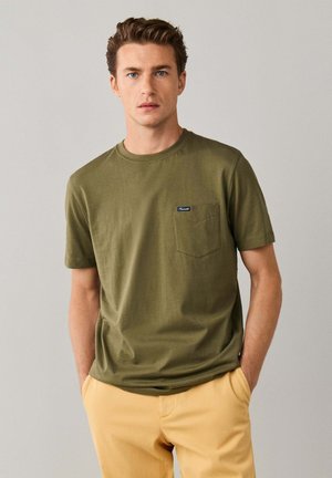 Joven con cabello corto y castaño, vistiendo una camiseta verde oliva y pantalones beige, de pie con las manos en los bolsillos contra un fondo liso.