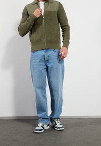 Olivgrön ribbad zip-up cardigan med en enfärgad övre panel, tillsammans med ljusblå löst sittande denimjeans och blågrå sneakers.