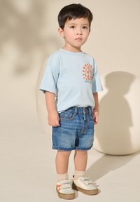 Mlad fant stoji, oblečen v svetlo modro majico z oranžnim grafičnim vzorcem, denim kratke hlače, bež nogavice in bele superge z oranžnimi poudarki.