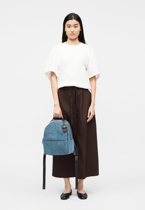 Kvinde stående med langt sort hår, iført en hvid kortærmet top, løse sorte bukser, sorte flade sko, og holder en blå rygsæk i hånden.