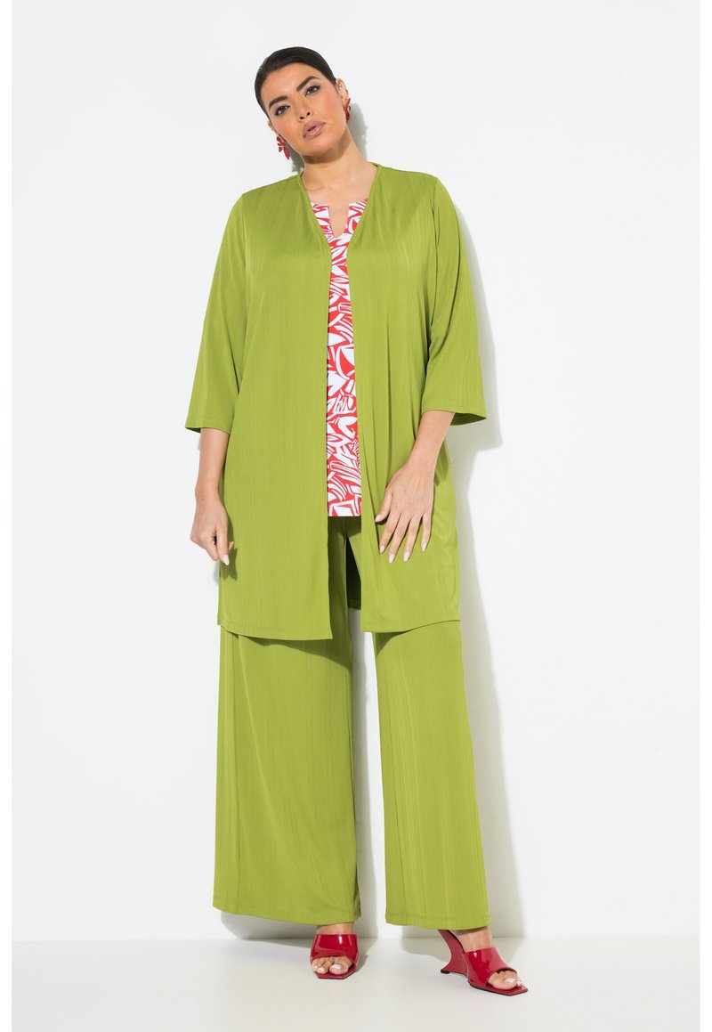 Zelený plisovaný kardigan přes červeno-bíle vzorovaný top a zelené kalhoty. Modelka má na sobě červené podpatky a náušnice se cvoky.