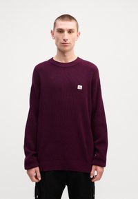 Oversized burgundy knit sweater with a textured pattern and ribbed crew neckline. Features a small white logo patch on the left chest.