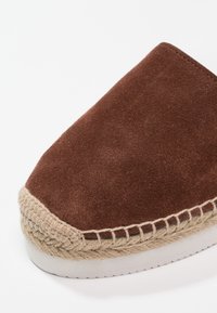 Mocassin en suédine brun avec un accent en jute tissé autour de la semelle, dotée d'un bout arrondi et d'une surface texturée. Semelle en caoutchouc blanche.