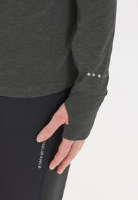 Person, die ein dunkelgraues Langarmoberteil mit Daumenloch und eine schwarze Leggings mit dem Text "ENDURANCE" an der Seite trägt.