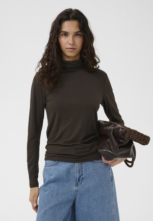 Žena s kučeravými tmavými vlasmi oblečená v tmavohnedom roláku a modrých džínsoch, držiaca veľkú hnedú textúrovanú tašku pod paží.