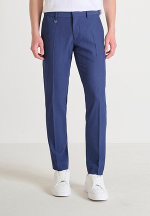 Blauwe broek van gestructureerde stof, met een platte voorkant, twee zijzakken en een slim fit. Draagt witte sneakers.