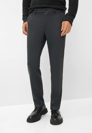 Man die donkergrijze op maat gemaakte broek en zwarte leren nette schoenen draagt, staand tegen een effen lichte achtergrond.