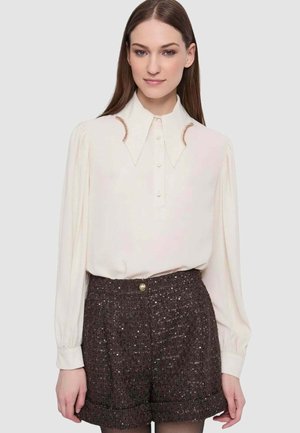 Blusa color crema a maniche lunghe con collo a punta e dettagli unici in tonalità rame, abbinata a pantaloncini marroni testurizzati con dettaglio a bottoni.