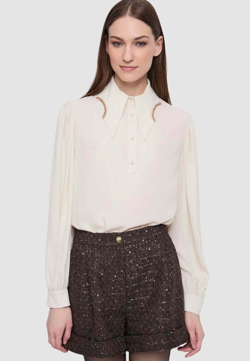 Blusa color crema a maniche lunghe con collo a punta e dettagli unici in tonalità rame, abbinata a pantaloncini marroni testurizzati con dettaglio a bottoni.