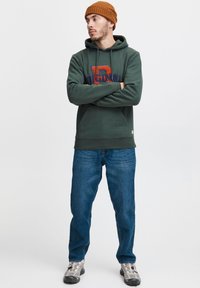 Tamsiai žalias džemperis su oranžiniais ir tamsiai mėlynais užrašais, derinamas su mėlynais džinsais ir pilkais batais. Modelis dėvi ruda ribota kepurę.