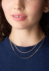 Collane a strati in oro e argento indossate sopra un maglione blu navy; una presenta delle perle rotonde lucenti, l'altra ha un design liscio e piatto.