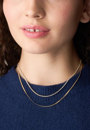 Des colliers en or et en argent superposés portés sur un pull bleu marine ; l'un présente des perles rondes brillantes, l'autre un design lisse et plat.