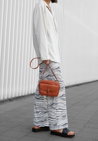 Camicia bianca a maniche lunghe, pantaloni larghi a fantasia con righe blu navy, sandali neri e una piccola borsa a tracolla in pelle marrone.