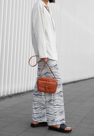 Camicia bianca a maniche lunghe, pantaloni larghi a fantasia con righe blu navy, sandali neri e una piccola borsa a tracolla in pelle marrone.