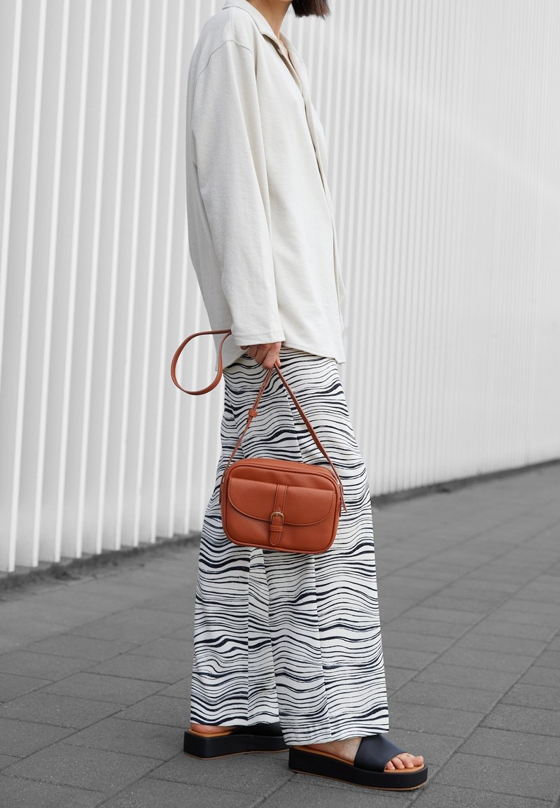 Camicia bianca a maniche lunghe, pantaloni larghi a fantasia con righe blu navy, sandali neri e una piccola borsa a tracolla in pelle marrone.