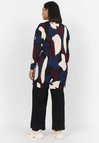 Tunique surdimensionnée en crème avec des motifs abstraits en marine, bordeaux et noir ; associée à un pantalon large noir et des baskets chunky beige.