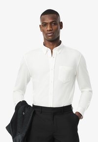 Weißes Langarmhemd mit Button-Down-Kragen, Brusttasche und leichter Textur, kombiniert mit schwarzen Hosen und einer dunklen Jacke.