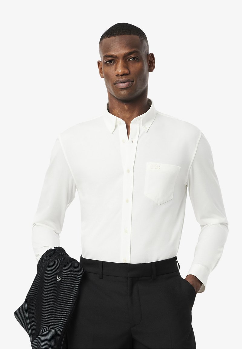 Weißes Langarmhemd mit Button-Down-Kragen, Brusttasche und leichter Textur, kombiniert mit schwarzen Hosen und einer dunklen Jacke.
