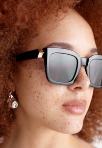 Óculos de sol oversized pretos com armações geométricas, lentes reflexivas e detalhe em dourado nas hastes. A pele apresenta sardas e textura natural.