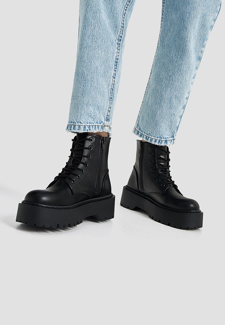 Bottes à plateforme épaisses noires avec lacets sur le devant et fermeture éclair latérale, portées avec un jean court bleu clair sur un fond uni.