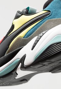 Kolorowy sneaker z czarną syntetyczną cholewką, żółtymi i morsko-zielonymi akcentami, teksturowanymi szarymi nakładkami oraz białą śródpodeszwą i czarną gumową podeszwą zewnętrzną.