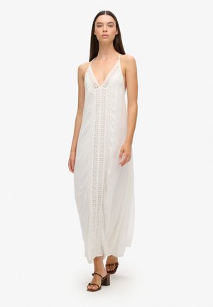Robe maxi blanche avec un décolleté en V profond, bordure en dentelle sur le devant et bretelles réglables. Modèle portant des sandales à talons marron.