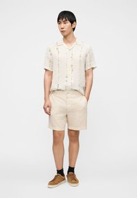 Λινό πουκάμισο με κοντά μανίκια και κεντήματα με λουλουδάτα σχέδια σε παστέλ χρώματα, σε συνδυασμό με σορτς σε ανοιχτό μπεζ και καφέ σουέντ αθλητικά παπούτσια.