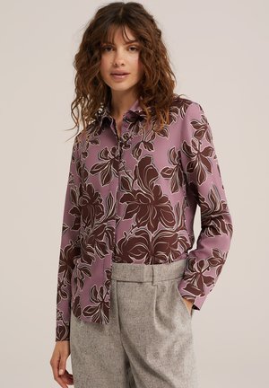 Femme portant un chemisier violet boutonné avec de grands motifs floraux marron et un pantalon taille haute gris clair.