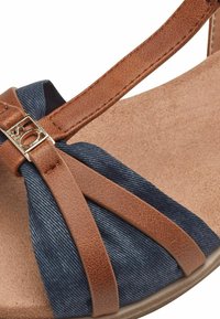 Cinghie in pelle sintetica marrone con un rivestimento in tessuto simile al denim blu, dotate di un dettaglio centrale a fiocco e una suola leggermente testurizzata.