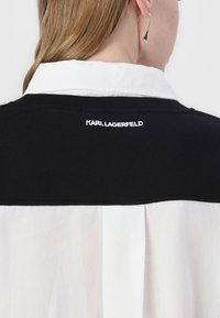 Svart och vit skjorta med en jämn textur; har en svart krage och bakpanel med "KARL LAGERFELD"-logotyp i vit brodering.
